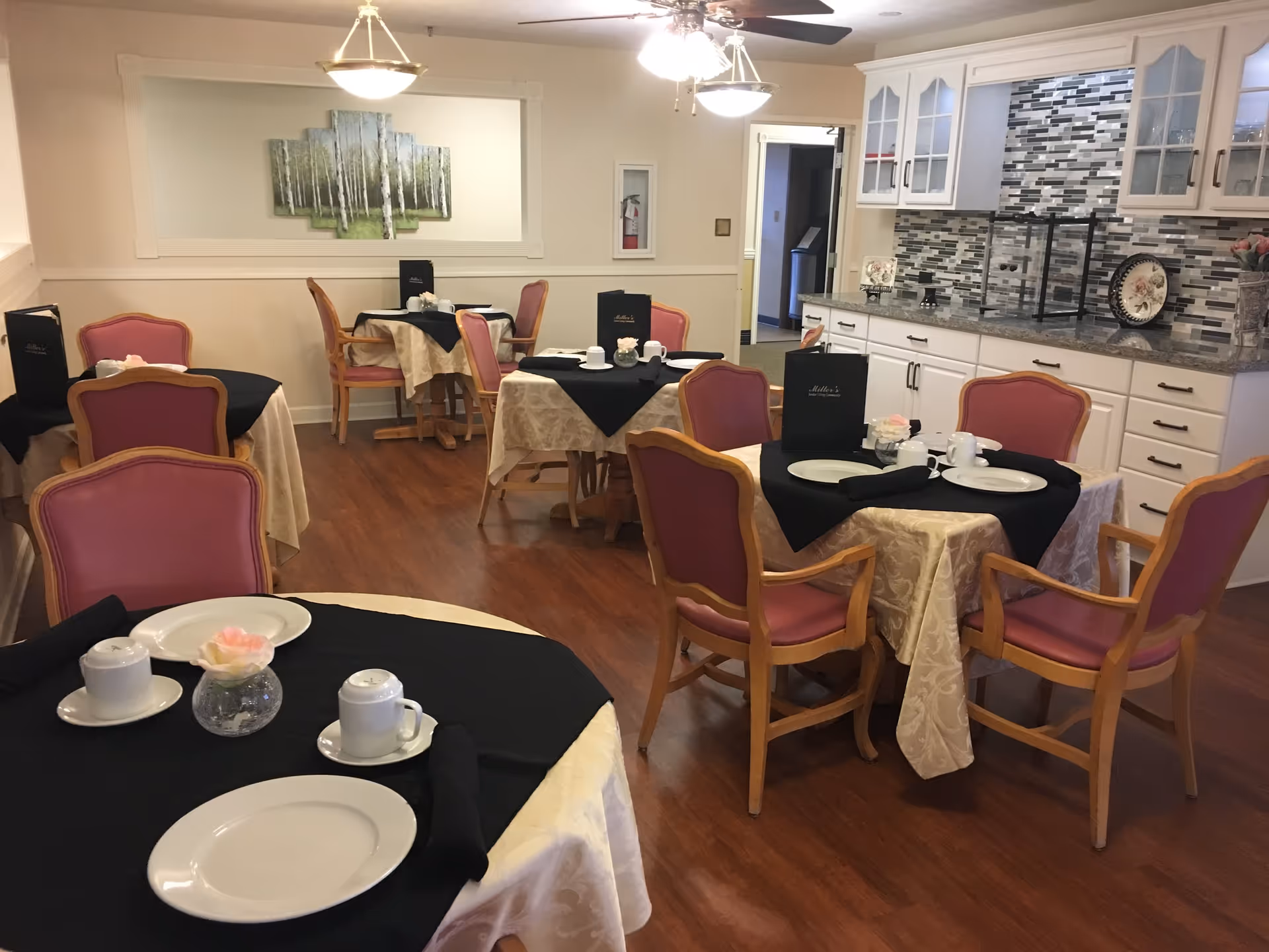 Dining room with several tables covered in beige tablecloths and black overlays, set with white plates, cups, and black napkins. Pink cushioned wooden chairs surround the tables. A decorative painting of birch trees hangs on the wall, and there is a cabinet with glass doors and a tiled backsplash on the right side.
