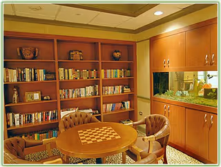 A cozy room with wooden bookshelves filled with books and decorative items, a round wooden table with a built-in chessboard, and four cushioned chairs arranged around the table. There is a cabinet with a glass display and plants inside on the right side of the room.