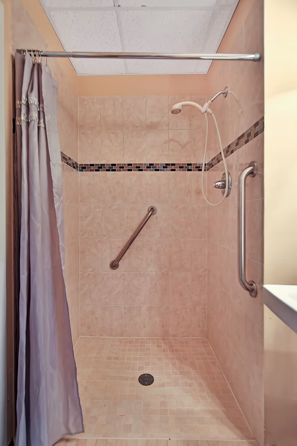A tiled walk-in shower with beige tiles and a decorative horizontal strip of small dark tiles. The shower has a handheld showerhead, two metal grab bars for support, and a purple shower curtain partially pulled to the side.