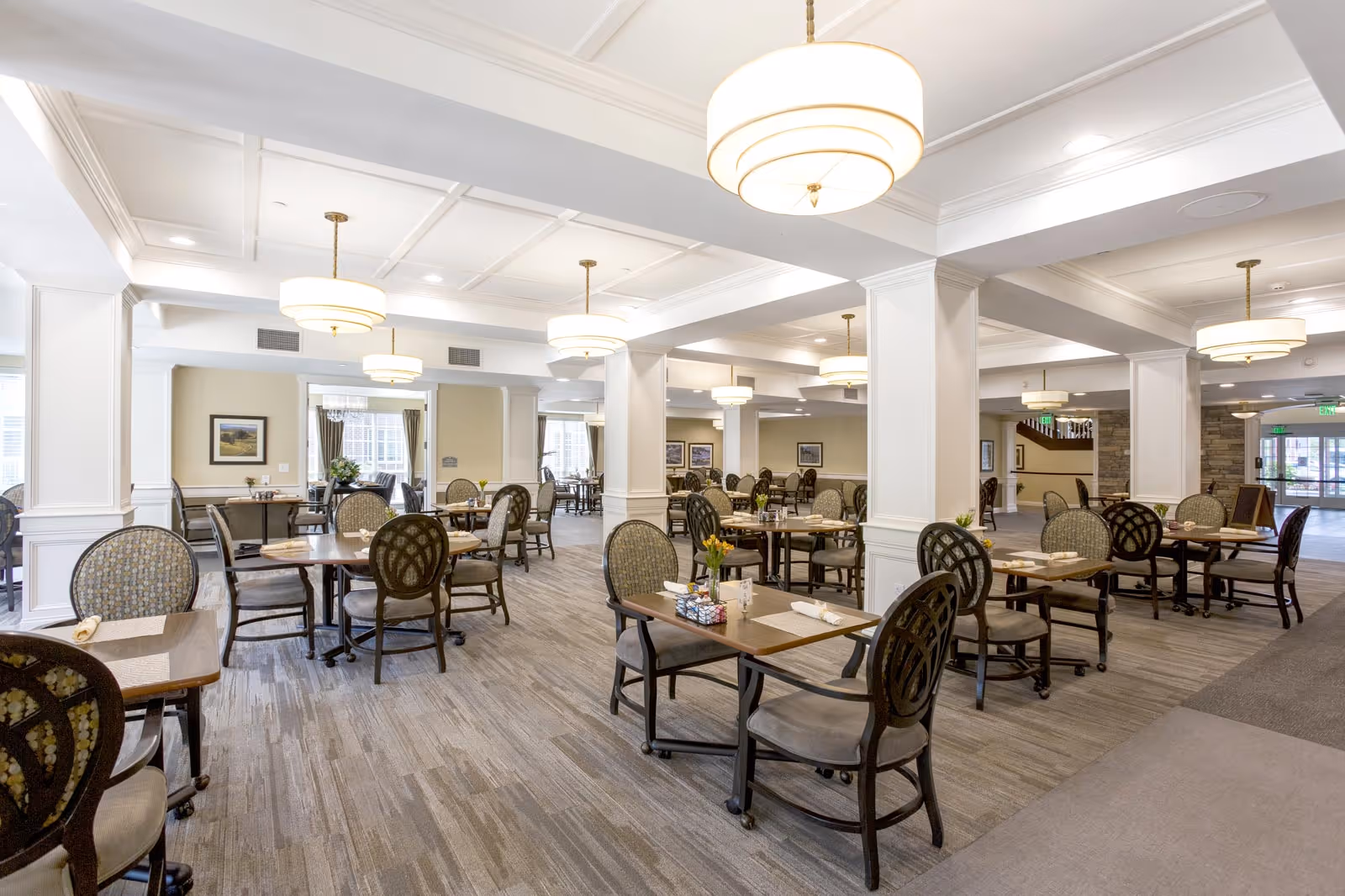 A spacious dining room with multiple tables and chairs arranged neatly. The room features white coffered ceilings with modern circular pendant lights hanging down. The floor is carpeted with a patterned design, and there are decorative columns throughout the space. The walls are light-colored with framed artwork, and there are windows allowing natural light to enter. Each table is set with placemats, napkins, and small flower arrangements.