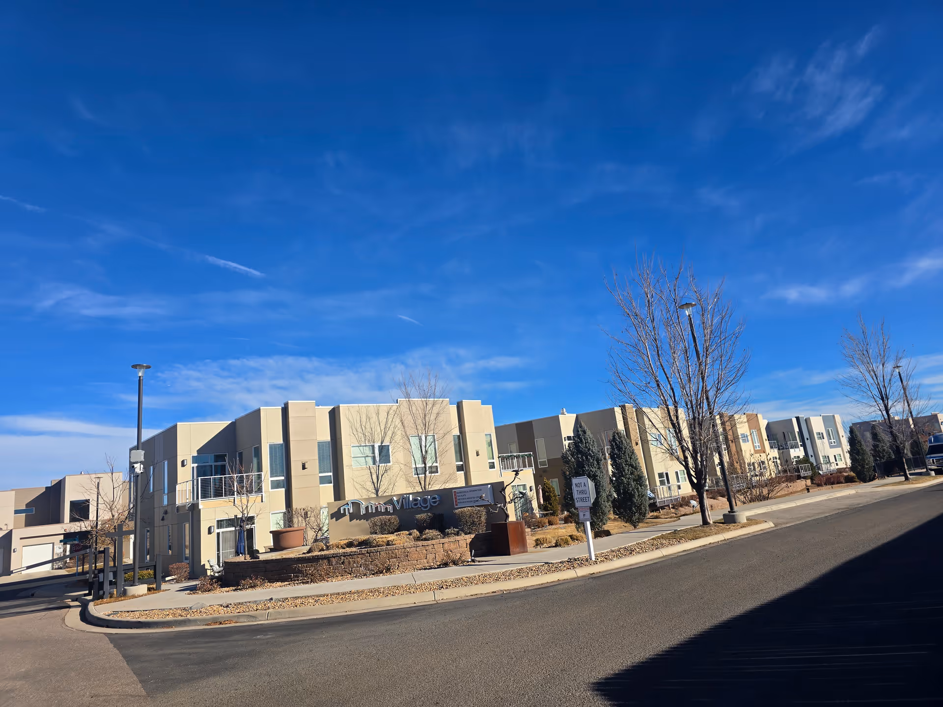 Front exterior of a low-rise senior living complex with beige modern buildings and bare trees under a bright blue sky.