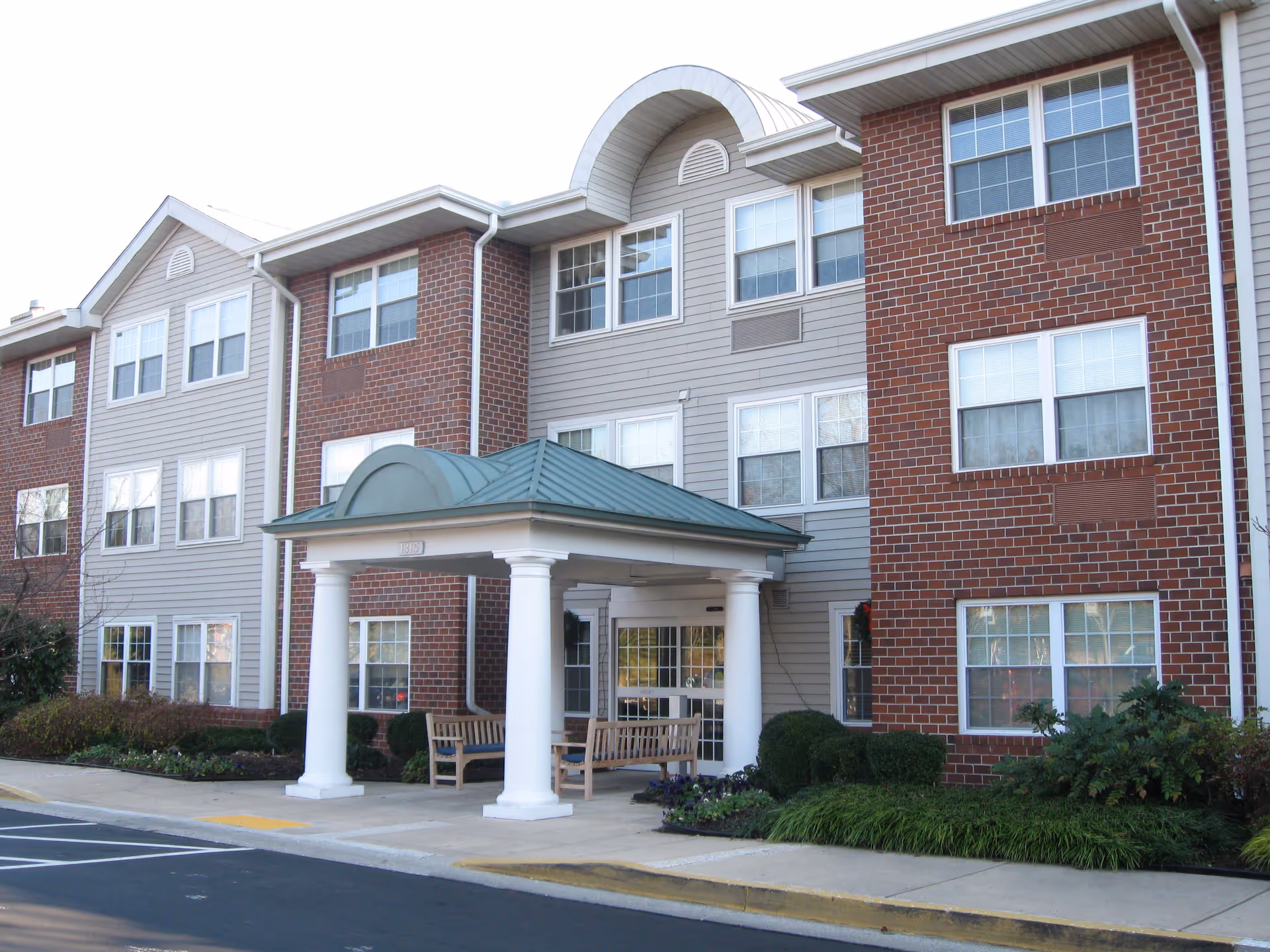 Exterior view of a senior living facility building with a covered entrance supported by white columns, benches under the entrance canopy, and landscaped bushes and plants along the sidewalk.