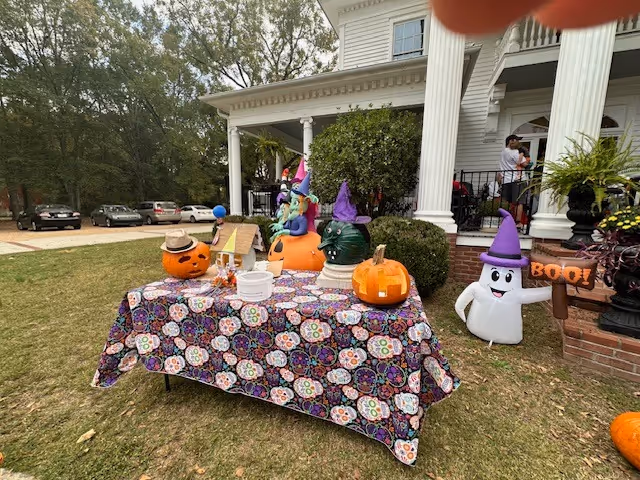 Front lawn and porch of a white house decorated for Halloween with a patterned table holding carved pumpkins and an inflatable ghost.