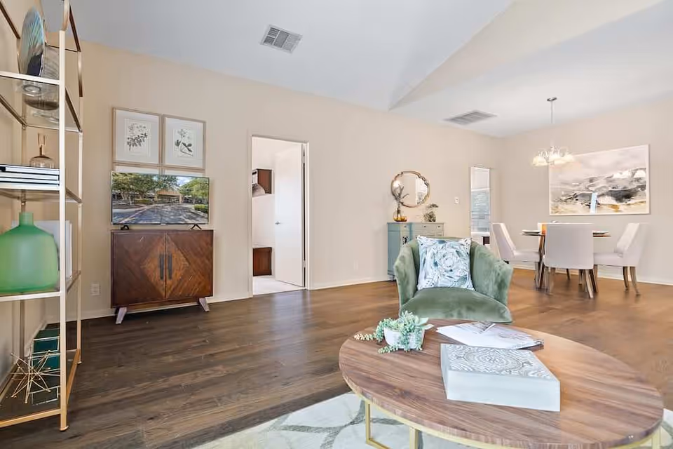 Open living and dining area with a green armchair and round wooden coffee table in the foreground, TV cabinet on the left and a dining table in the background.