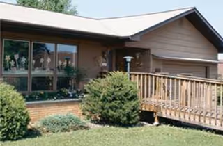 Single-story brick building with large windows, a wooden deck, and manicured shrubs.
