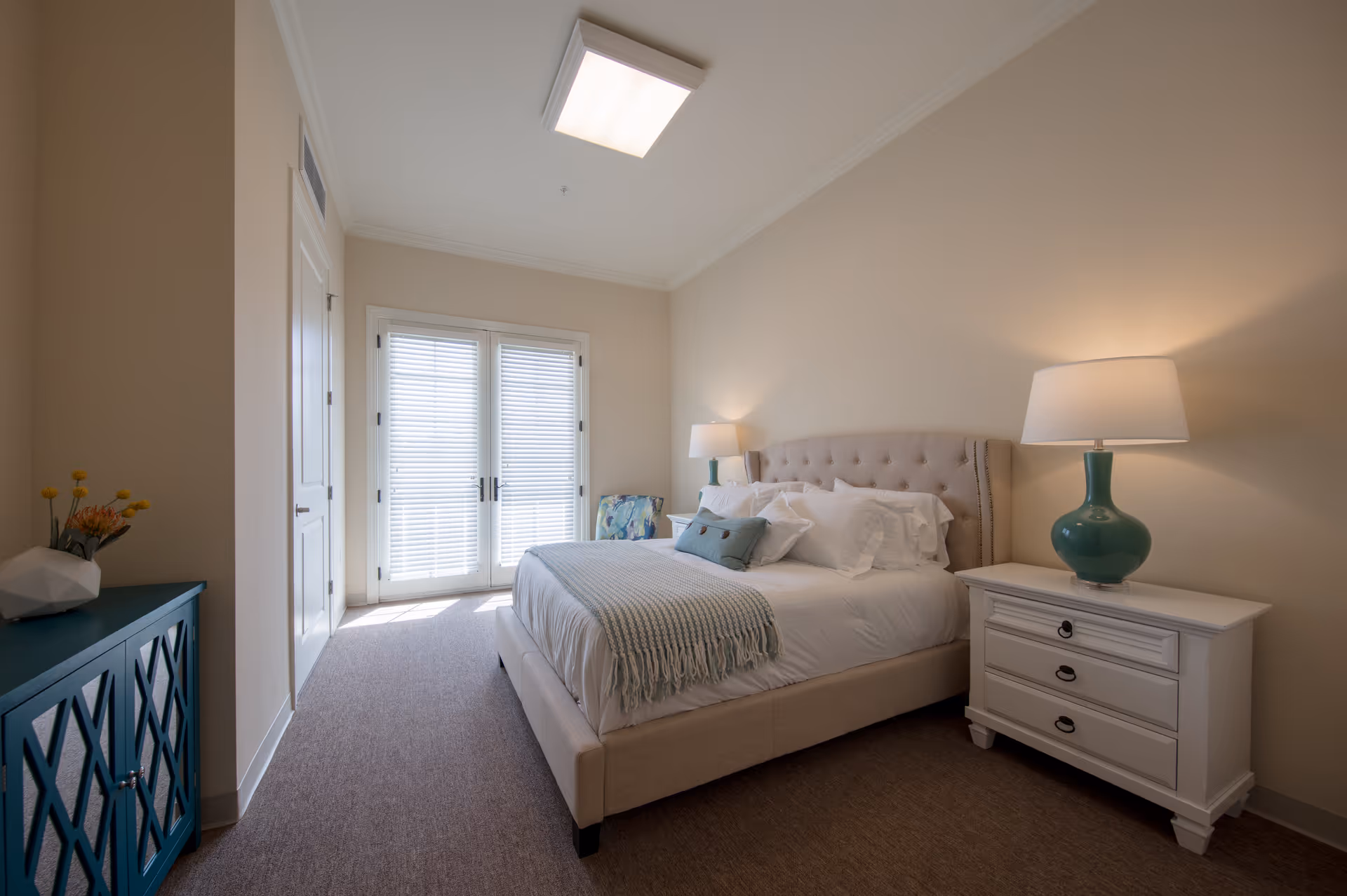 Well-lit bedroom with an upholstered bed, white nightstand and lamps, French doors with blinds, and a teal cabinet.