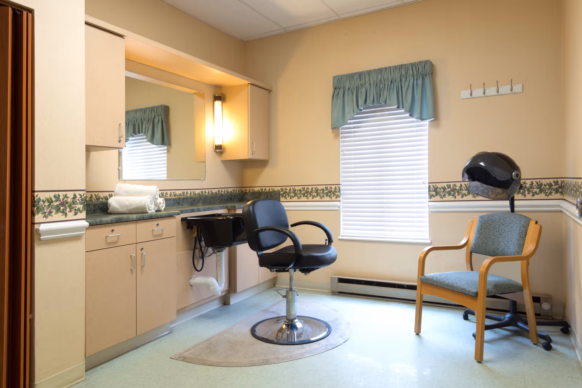 A small salon room with a black salon chair in front of a sink and countertop with cabinets. There are two rolled white towels on the counter. A window with closed blinds and a green valance is on the wall. Next to the window is a wooden chair with blue upholstery and a black hair dryer hood attached to a stand. The walls have a floral wallpaper border and the floor is light-colored.