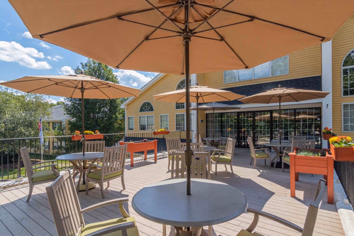 Outdoor patio area at The Village at East Farms with round tables and chairs under large beige umbrellas. The patio is surrounded by a railing with flower boxes and overlooks greenery and other buildings. The building exterior is yellow with large windows and a glass door.