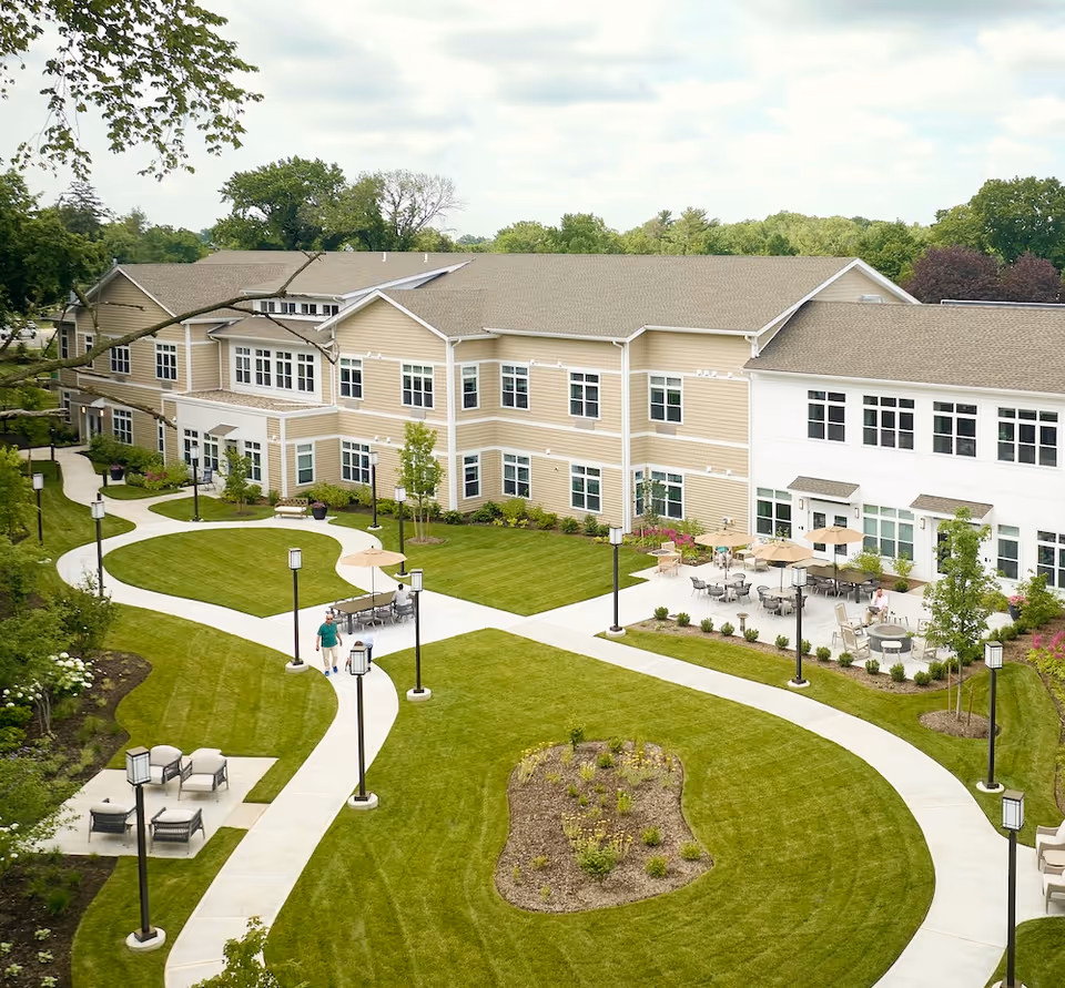 Exterior view of Monarch Coopers Corner facility showing a two-story beige and white building with many windows, surrounded by well-maintained green lawns, paved walkways, outdoor seating areas with tables and umbrellas, and several lamp posts. A person is walking on one of the paths.