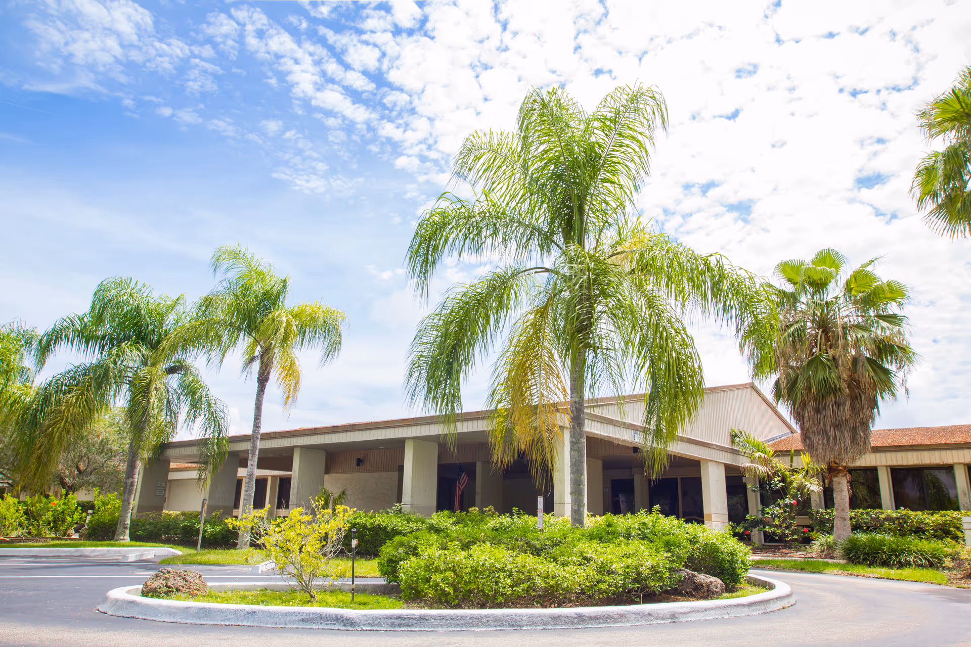Exterior view of Brookwood Gardens Nursing & Rehabilitation Center showing a single-story building with a covered entrance, surrounded by palm trees and green shrubs under a partly cloudy sky.