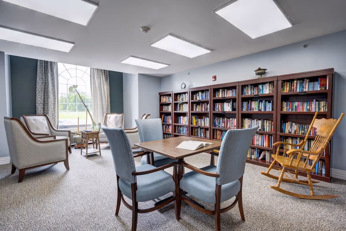 A cozy library room with a large window covered by curtains, several bookshelves filled with books, a wooden rocking chair, a table with four upholstered chairs, and two armchairs near the window.