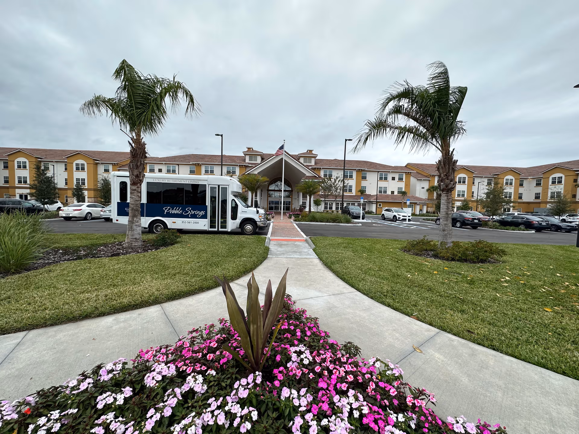 Front exterior view of Pebble Springs Retirement Community building with a driveway, parking lot, palm trees, and a shuttle bus parked near the entrance. There is a flower bed with pink and white flowers in the foreground and an American flag above the entrance.