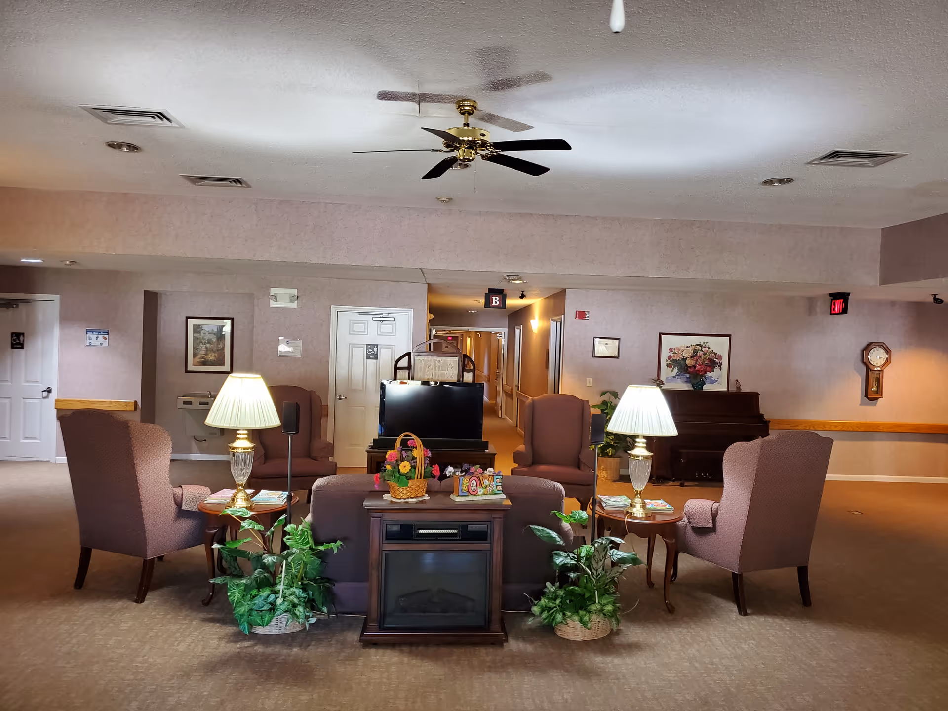 Common lounge area with armchairs arranged around a TV and lamps, a piano against the wall, plants, and a ceiling fan.