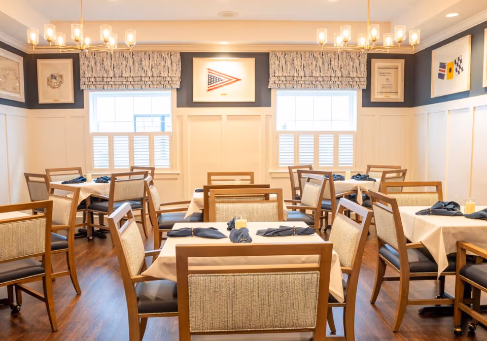 A bright dining room with several tables covered in white tablecloths, each set with dark cloth napkins and napkin rings. The room has wooden chairs with cushioned backs and seats, hardwood flooring, two windows with patterned valances and white shutters, and framed nautical-themed artwork on the walls.