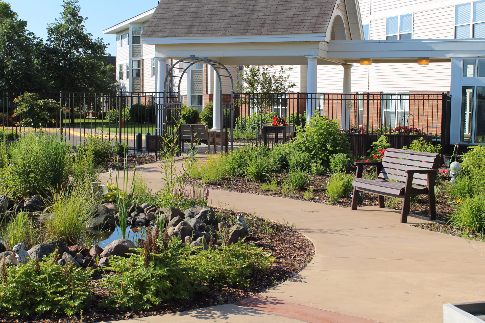 A landscaped outdoor garden area at Oak Meadows Senior Living featuring a curved concrete pathway, a wooden bench, various green plants and shrubs, a small pond with rocks, and a white building with a covered porch in the background.