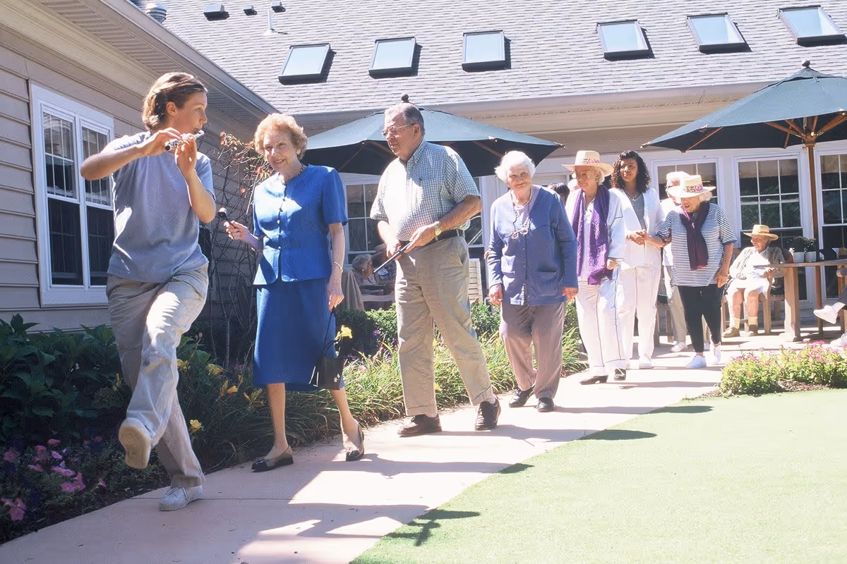 A group of elderly people walking in a line outdoors along a paved path next to a building with large windows and umbrellas. A younger person is leading the group, appearing to play a flute or similar instrument. The setting is sunny with greenery and flowers along the path.