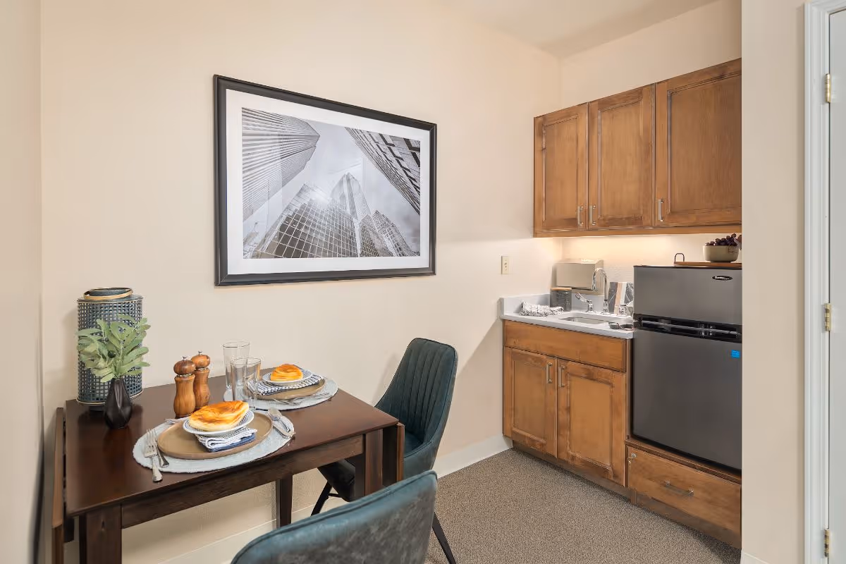A small dining area with a dark wooden table set for two, featuring plates with pastries, glasses, and cutlery. Two dark green upholstered chairs are positioned at the table. To the right, there is a kitchenette with wooden cabinets, a small sink, a toaster, and a compact refrigerator. A framed black and white photo of tall skyscrapers is hung on the beige wall above the dining table.