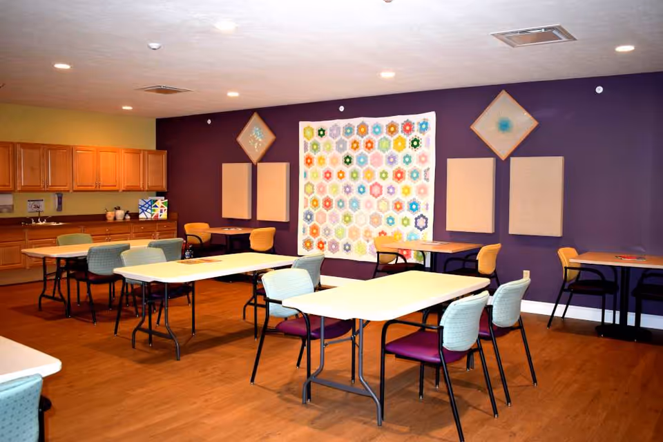 A bright room with several tables and chairs arranged for group activities or dining. The walls are painted purple and green, with a colorful quilt hanging on the purple wall. Wooden cabinets and a countertop with a sink are visible along one wall. The floor is wooden, and the ceiling has recessed lighting.