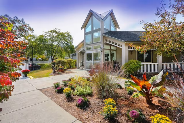 Exterior view of a senior living facility building with large windows and a peaked roof. The building is surrounded by a well-maintained garden with colorful flowers and shrubs. A concrete walkway leads to the entrance, and trees with autumn foliage are visible around the property.