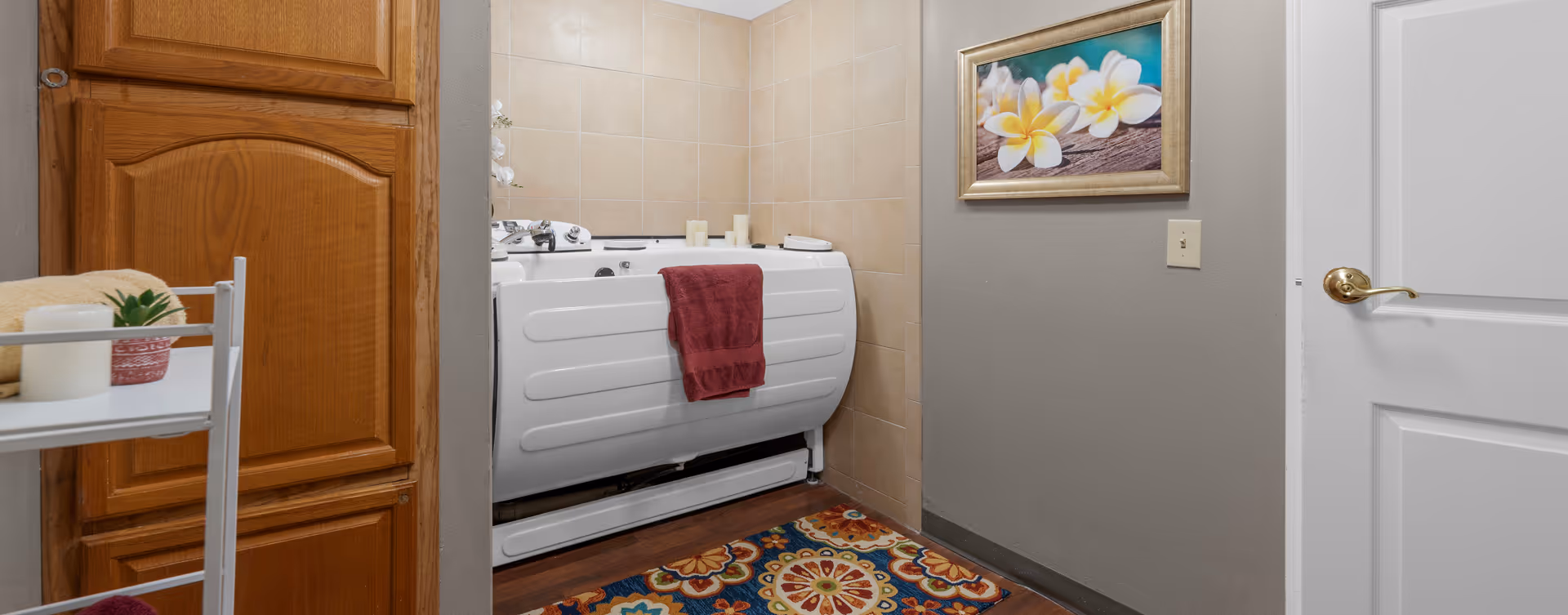 A bathroom corner featuring a white walk-in bathtub with a maroon towel hanging on its side. The walls are tiled in beige, and there is a colorful patterned rug on the wooden floor. To the left, there is a wooden cabinet and a white metal rack holding a candle, a small plant, and a folded towel. On the right wall, a framed picture of white and yellow flowers is hung above a light switch, next to a white door with a brass handle.