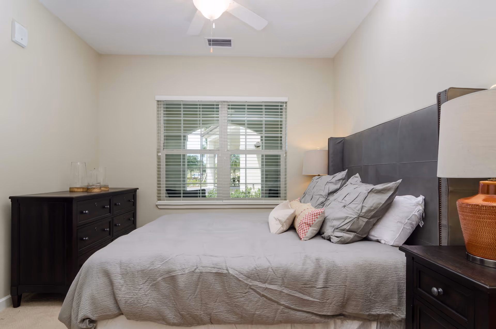 A bedroom with a large bed covered in a gray bedspread and multiple pillows. There is a dark wooden dresser with glass containers on top on the left side and a matching nightstand with an orange lamp on the right side. A window with white blinds is centered on the back wall, and a ceiling fan is visible above.