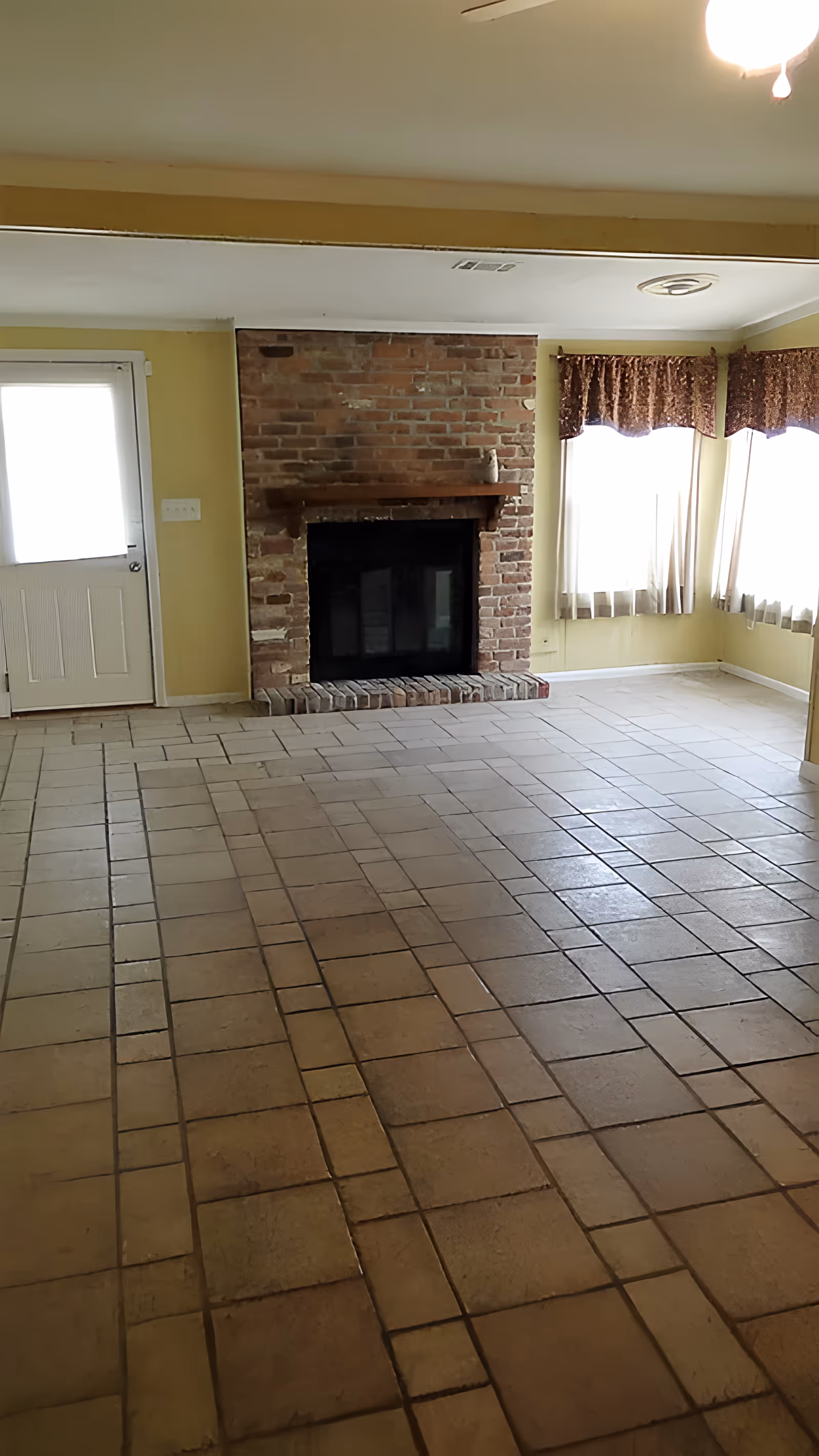 An empty room with a tiled floor, a brick fireplace in the center of the far wall, a door to the left, and two windows with curtains on the right side. The walls are painted yellow and the ceiling is white with a light fixture.