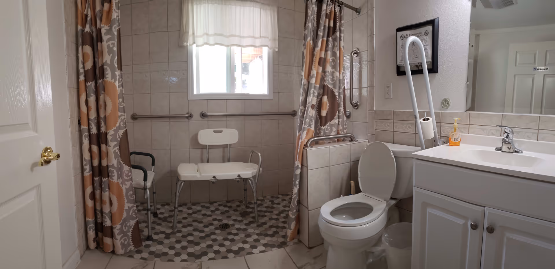 A bathroom with a walk-in shower area featuring a white shower chair and grab bars. The shower has a patterned curtain with orange and gray circular designs. There is a window with a white curtain above the shower. Next to the shower is a toilet with a grab bar and a white vanity with a sink, faucet, and a bottle of hand soap. The walls are tiled and there is a framed picture hanging above the toilet.