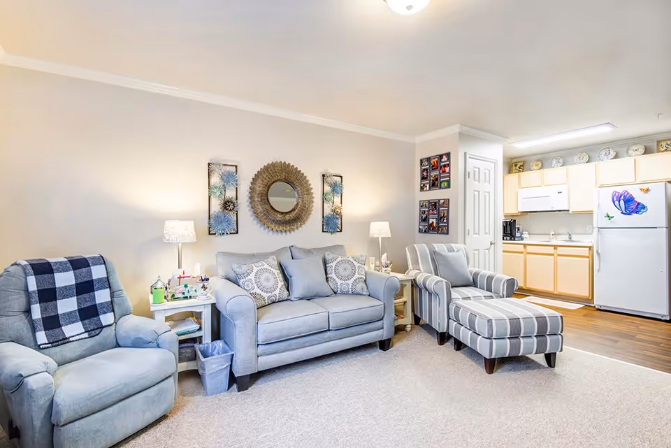 A cozy living room area with a light blue armchair draped with a checkered blanket, a matching blue sofa with decorative pillows, and a striped armchair with an ottoman. The walls are decorated with a round mirror and floral wall art. Two side tables with lamps flank the sofa. In the background, there is a small kitchen with light wood cabinets, a white microwave, and a refrigerator decorated with butterfly magnets.