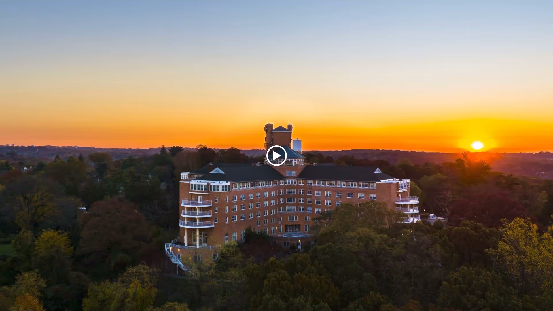 Aerial view of a large brick senior living building on a wooded hill at sunset.