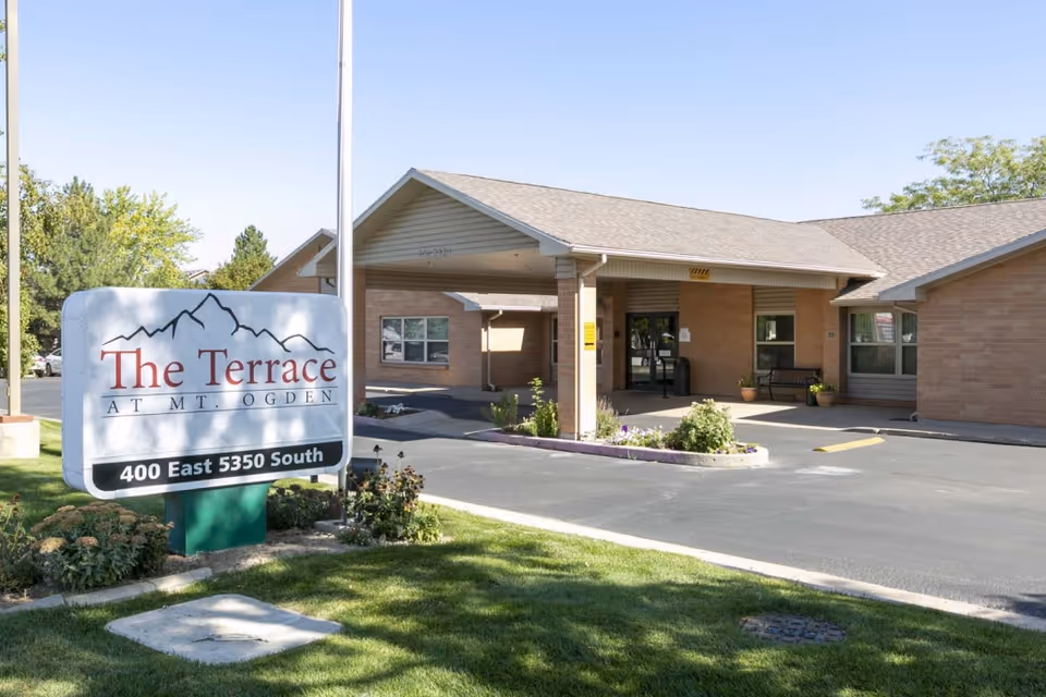 Exterior view of The Terrace at Mt. Ogden senior living facility showing the entrance with a covered drop-off area, a sign with the facility name and address, and surrounding greenery under a clear sky.