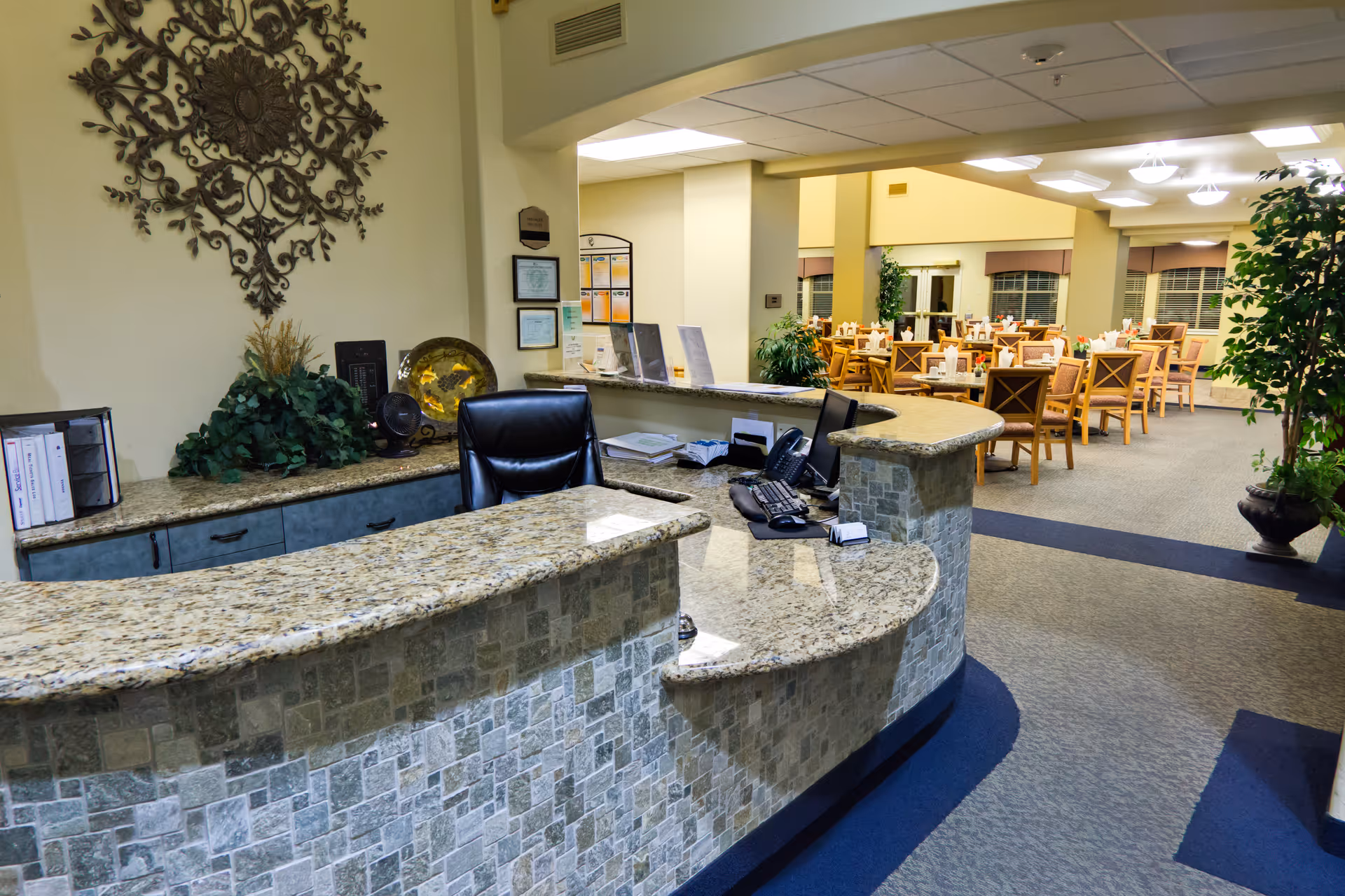 Reception desk area with a curved granite countertop and a black office chair. Behind the desk, there is a decorative wall piece, some plants, and office supplies. In the background, there is a dining area with multiple wooden tables and chairs set up, illuminated by ceiling lights.