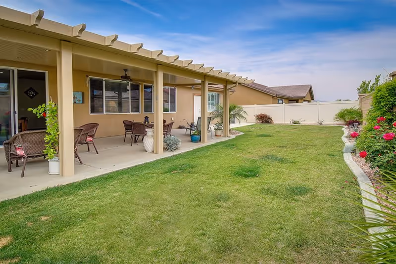 Covered patio with wicker seating and potted plants overlooking a grassy fenced backyard with flower beds.