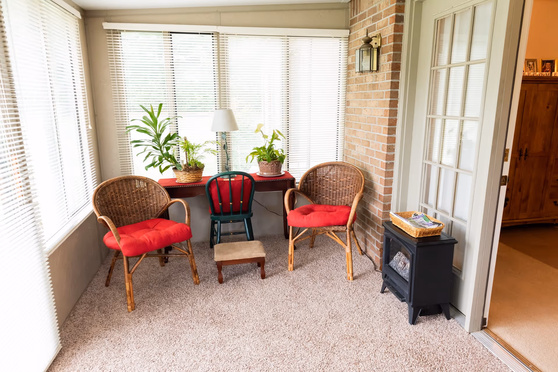 A cozy sunroom with large windows covered by white blinds. The room features two wicker chairs with red cushions, a small green chair, a small footstool, a table with a red cloth holding three potted plants and a lamp, a small black electric fireplace with a basket on top, and a brick accent wall. A glass door leads to an adjacent room with wooden furniture.