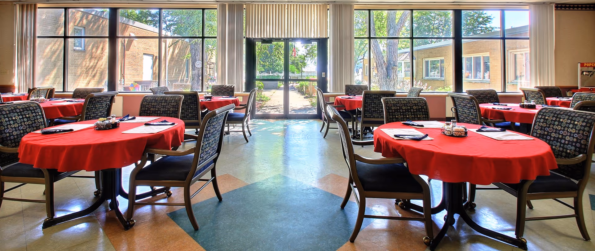Communal dining room with round tables draped in red tablecloths and chairs arranged before large windows and a glass exit door.
