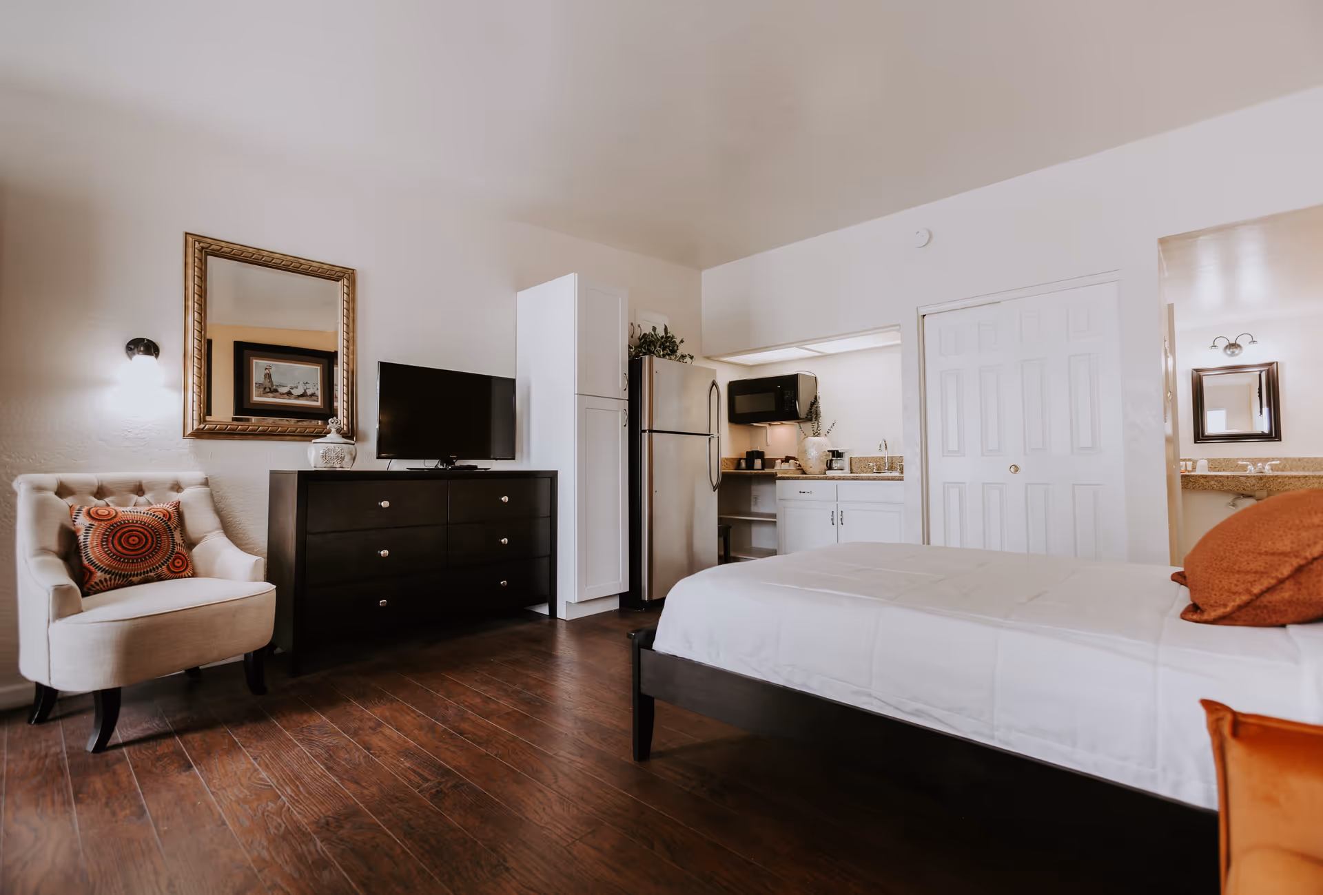 A cozy studio apartment interior featuring a bed with white bedding and orange pillows, a dark wood dresser with a flat-screen TV on top, a beige armchair with a decorative pillow, a large mirror on the wall, and a small kitchenette with a refrigerator, microwave, and sink. The room has wooden flooring and light-colored walls.