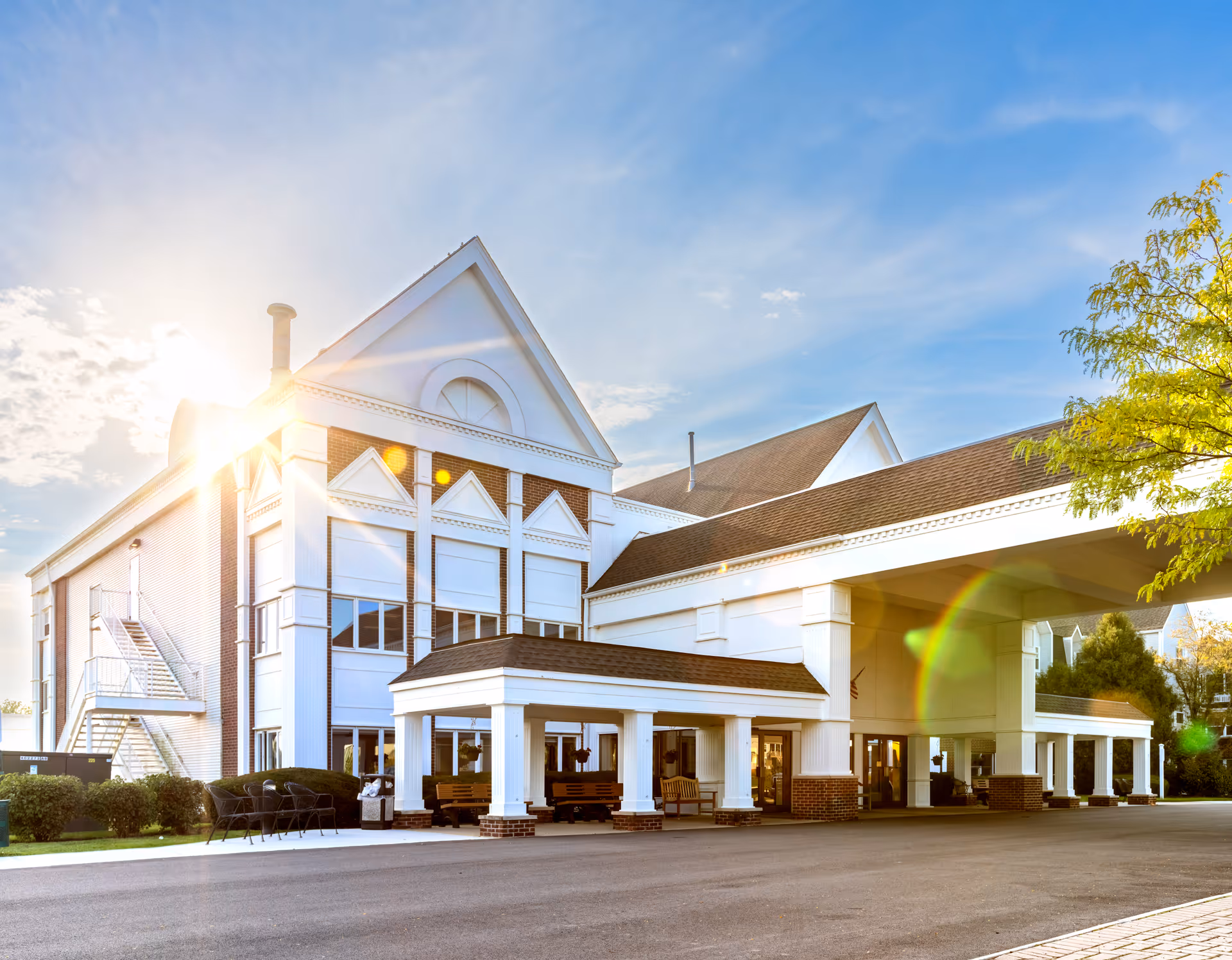Sunlit front exterior of a large white multi-story senior living building with a covered entry portico and benches.