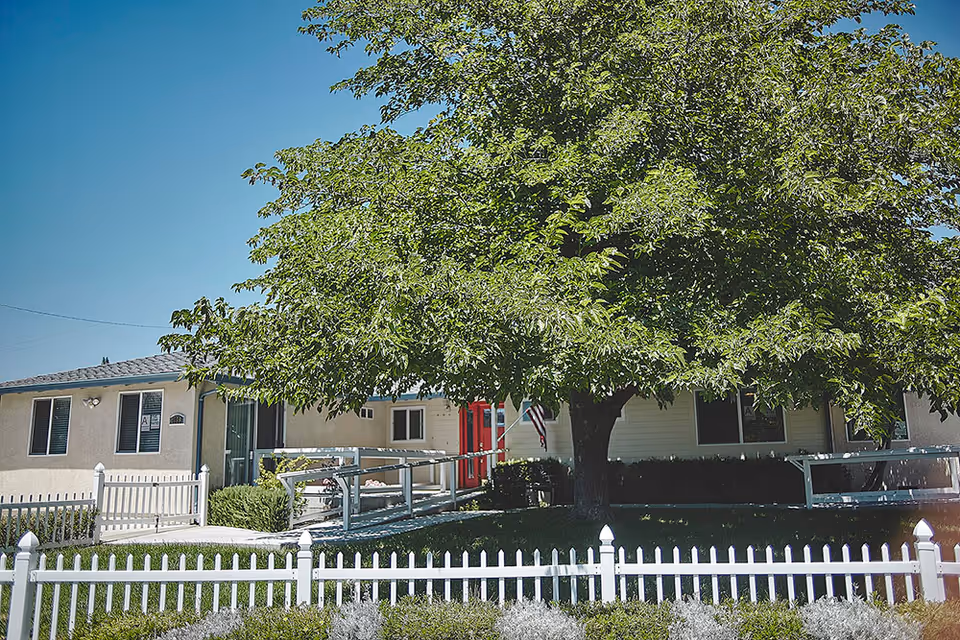 Exterior view of Abigail's Beckham Care Home showing a single-story building with beige walls and a red door. There is a large leafy tree in front of the building, a white picket fence, and a wheelchair ramp leading to the entrance under a clear blue sky.