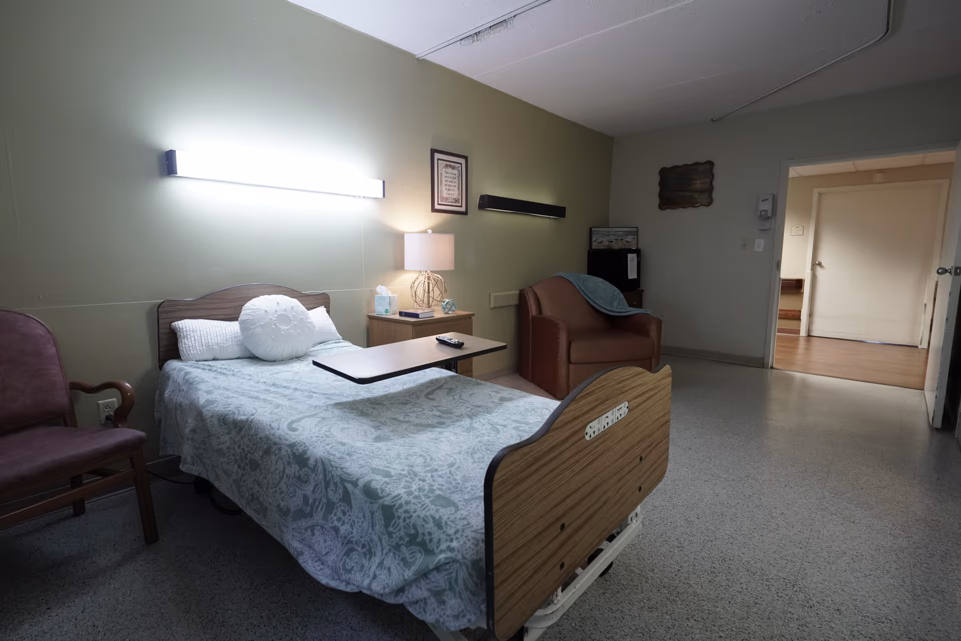 A simple healthcare facility room with a single hospital bed covered with a patterned bedspread, a small overbed table, a wooden nightstand with a lamp and tissues, a brown armchair with a blue blanket draped over it, and a purple chair. The walls are painted light green and gray, with framed pictures hanging. The floor is a speckled linoleum, and there is a doorway leading to another room.