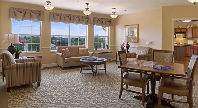 Sunlit common lounge with sofas, armchairs, a round coffee table and a dining table near large windows overlooking greenery.