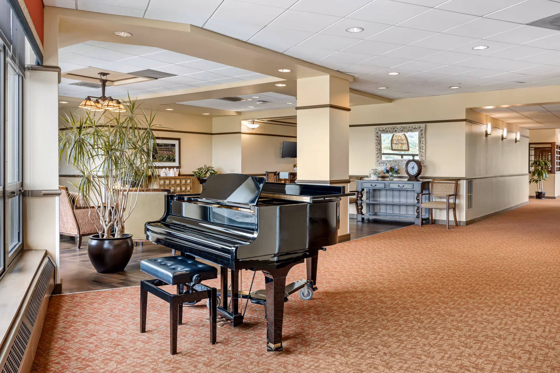 Interior view of a senior living facility lounge area featuring a black grand piano with a matching bench on a patterned carpet. To the left, there is a large potted plant near windows, and in the background, there are seating areas with chairs and tables, a decorative console table with flowers and a clock, and soft lighting fixtures on the walls and ceiling.