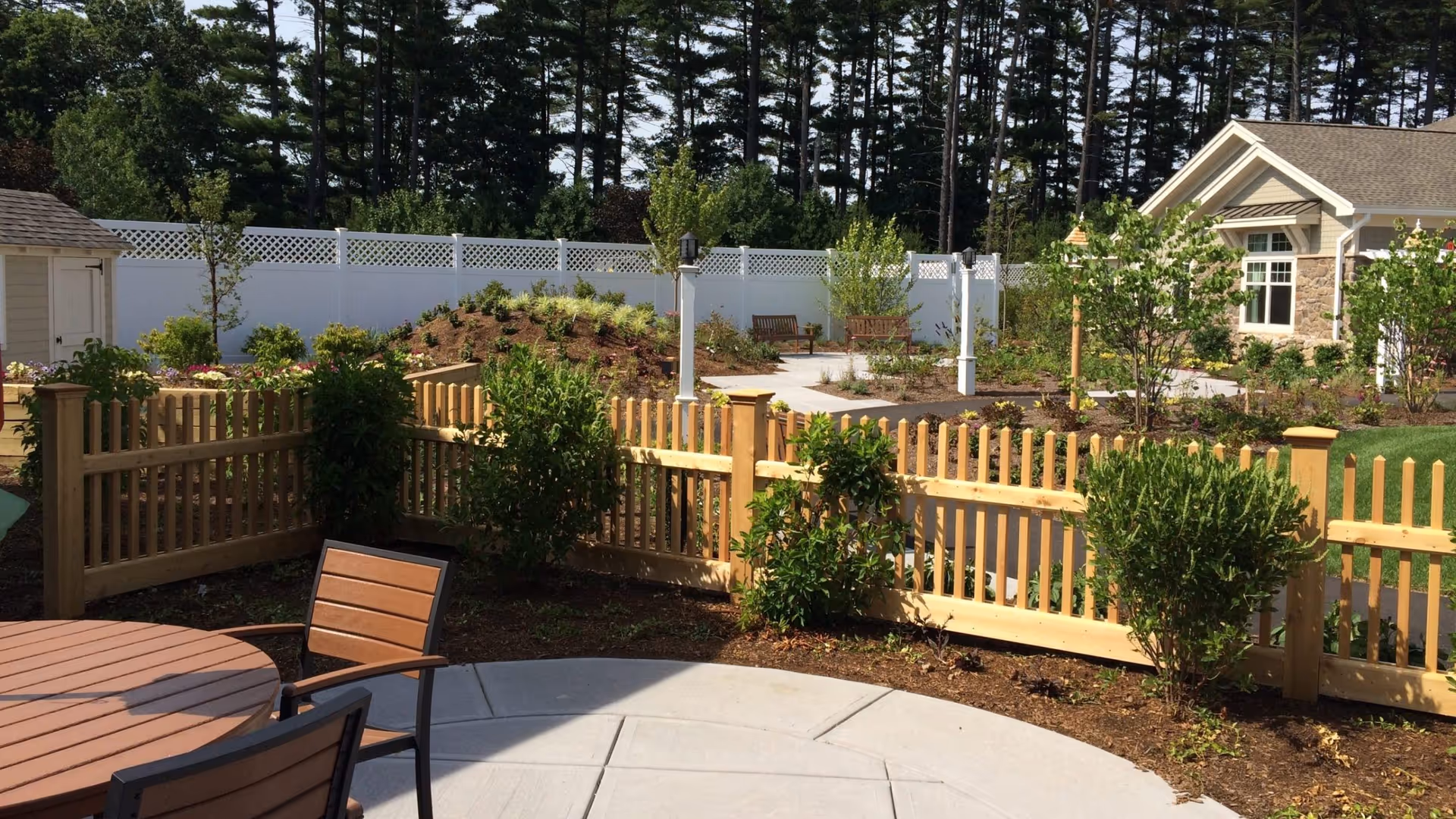 Outdoor patio area with a round wooden table and chairs, surrounded by a wooden picket fence and landscaped garden beds with shrubs and small trees. In the background, there is a paved walkway, benches, a white privacy fence, and a building with stone and siding exterior. Tall pine trees are visible beyond the fence.