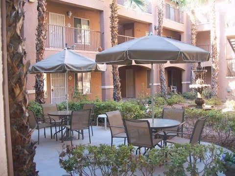 Outdoor courtyard area of Court Senior Apartments featuring round tables with umbrellas and chairs, surrounded by palm trees and shrubbery, with apartment balconies and doors visible in the background.