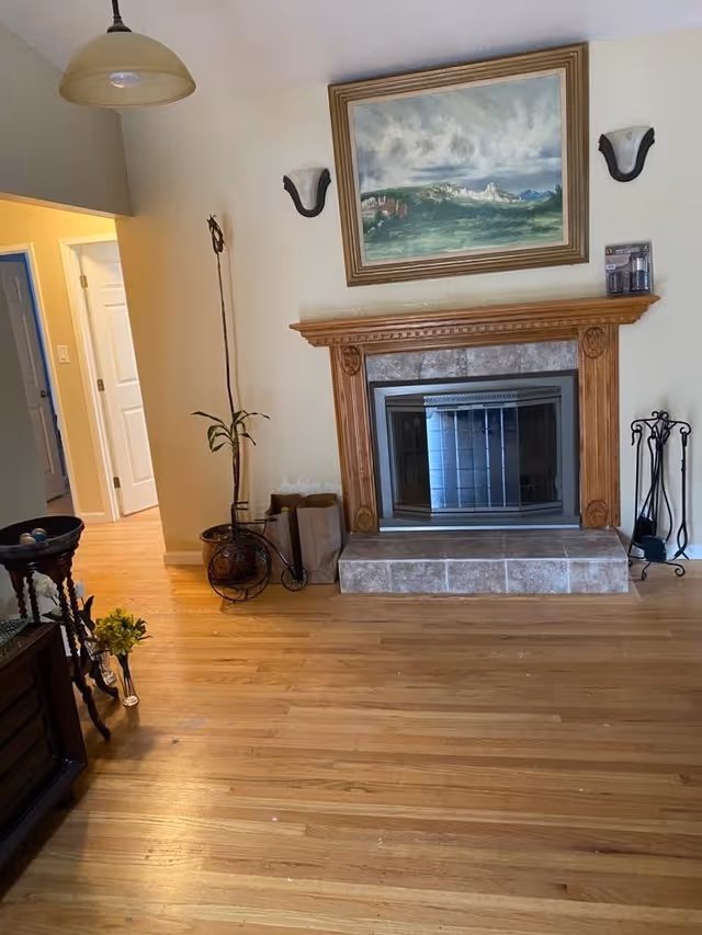 A cozy living room with a wooden floor, a fireplace with a wooden mantel and tiled hearth, a framed landscape painting above the fireplace, two wall sconces on either side of the painting, a tall plant in a decorative pot, a small table with a vase of yellow flowers, and an open doorway leading to another room.