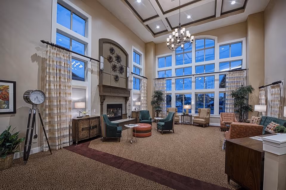 A spacious and elegant living room with high ceilings and large windows letting in natural light. The room features a fireplace with decorative art above it, multiple comfortable chairs arranged around small tables, a large chandelier hanging from the ceiling, and patterned curtains on the windows. There are also plants and lamps adding to the cozy atmosphere.