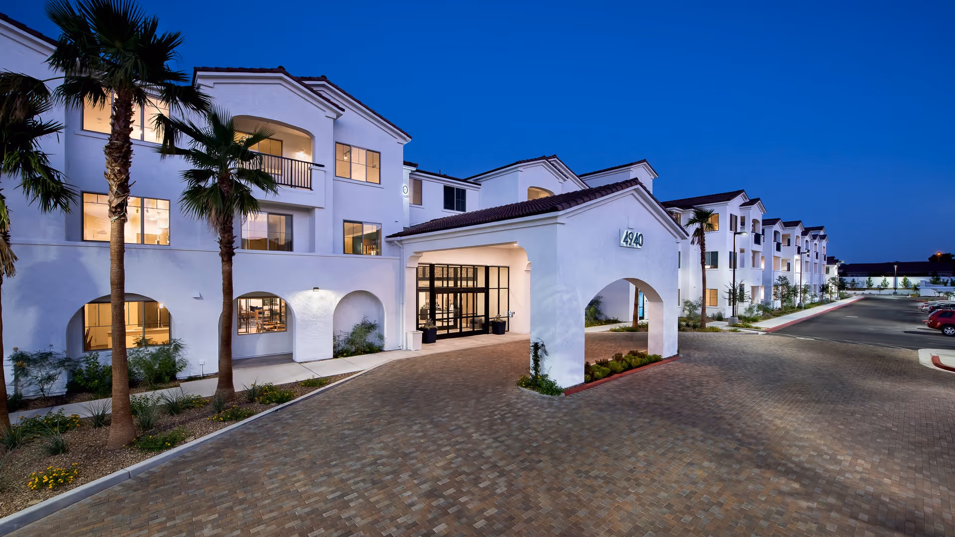 Exterior view of a modern three-story senior living facility named Revel Vegas at dusk, featuring white stucco walls, arched entryways, palm trees, and a paved driveway with parked cars.