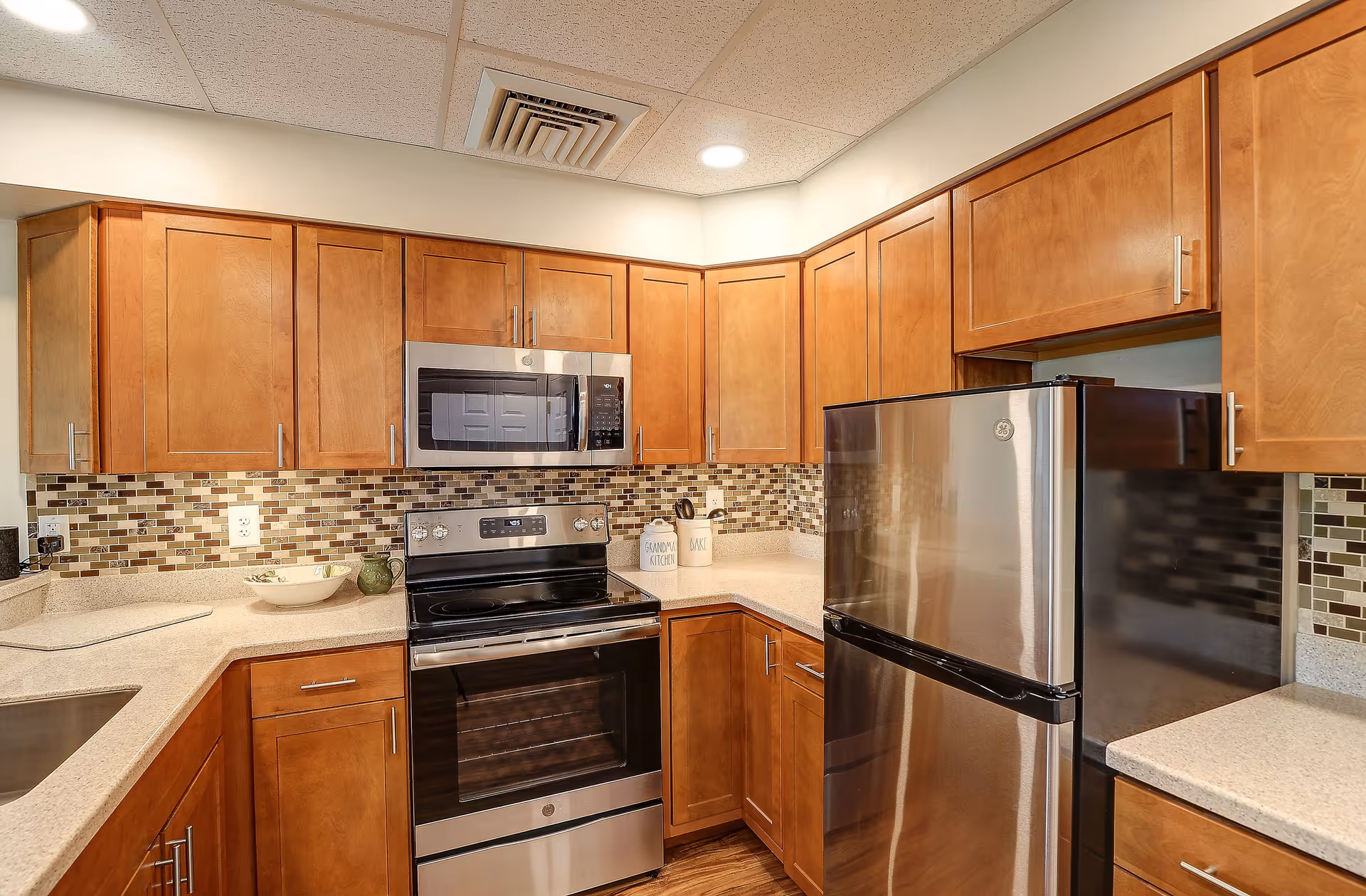 A modern kitchen with wooden cabinets, a stainless steel refrigerator, stove, and microwave. The backsplash features a mosaic tile design in neutral tones, and the countertops are light-colored. The kitchen has recessed lighting and a clean, organized appearance.