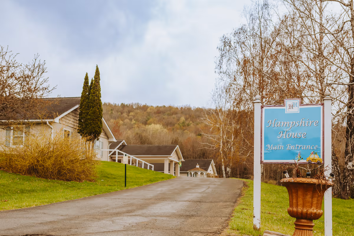Driveway leading to Hampshire House senior living buildings with a blue 'Hampshire House Main Entrance' sign in the foreground.