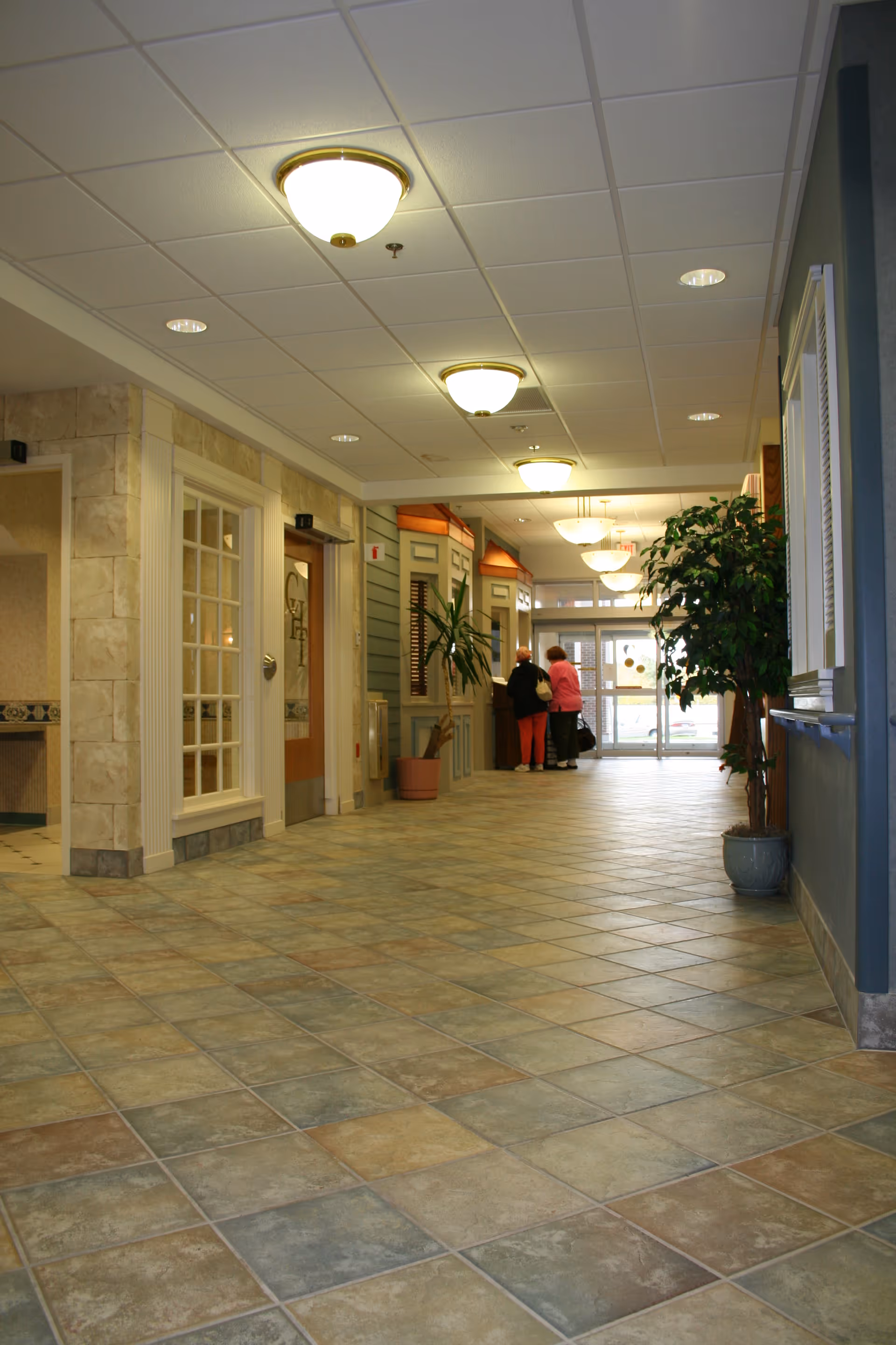 A wide, tiled hallway in a nursing home or rehabilitation center with ceiling lights, potted plants, and two people standing near the entrance door at the far end.