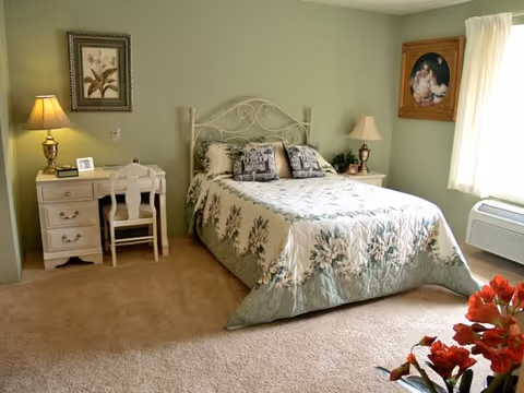 A cozy bedroom with a double bed covered in a floral quilt, two decorative pillows, a white metal headboard, a white desk with a chair and a lamp on the left side, a nightstand with a lamp and a plant on the right side, a framed floral picture on the wall above the desk, a framed portrait on the adjacent wall, beige carpet flooring, and a window with sheer curtains letting in natural light.