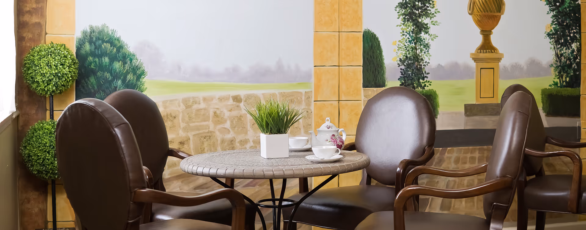 A round table with a decorative pattern on the surface surrounded by four brown leather chairs with wooden armrests. On the table, there is a small white square pot with green grass-like plant, a white teapot with floral design, and two white teacups with saucers. The background features a mural painting of a garden scene with greenery, stone walls, and a large urn on a pedestal.